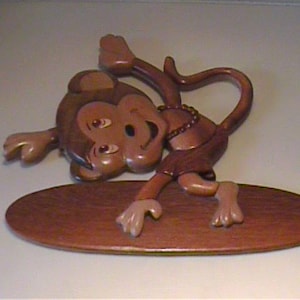 May include: A wooden monkey figurine in a surfing pose. The monkey is brown with a tan face and hands, wearing a brown loincloth and a necklace. It is positioned on a brown surfboard-shaped base.