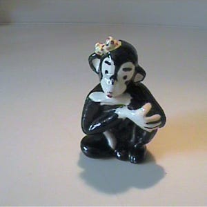 May include: A small, ceramic monkey figurine in a seated position. The monkey is primarily black with white facial features and hands. It wears a small crown. The figurine has a glossy finish and is set against a plain background.