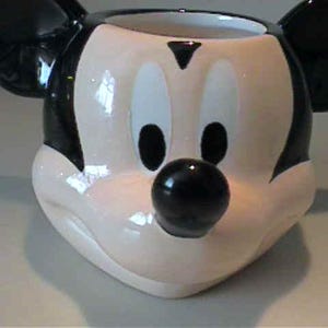 May include: A ceramic mug shaped like the head of Mickey Mouse, featuring a glossy finish in black, white, and flesh-toned colors. The mug displays the character's iconic large round ears, oval eyes, a small triangular brow, and a prominent black nose.