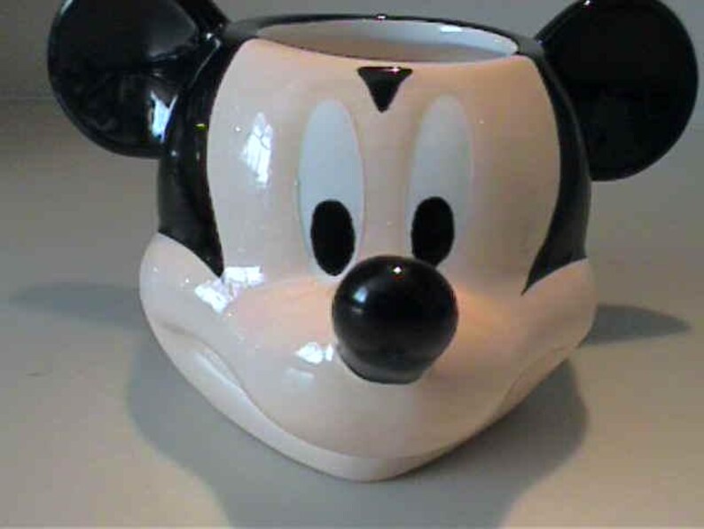 May include: A ceramic mug shaped like the head of Mickey Mouse, featuring a glossy finish in black, white, and flesh-toned colors. The mug displays the character's iconic large round ears, oval eyes, a small triangular brow, and a prominent black nose.