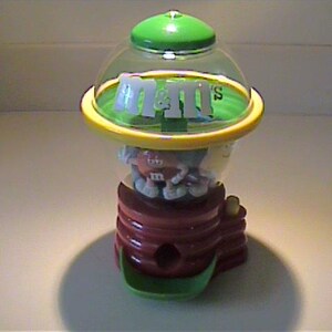 May include: A plastic M&M's candy dispenser featuring a clear dome top with a green lid and yellow rim. The base is brown and cylindrical with a green dispensing tray at the bottom. Inside the clear dome, a small M&M character figure is visible.