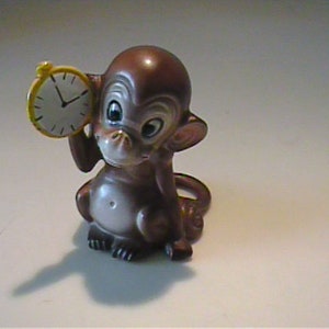 May include: A brown monkey figurine holding a yellow clock with black hands. The monkey has large eyes and a white belly. The clock shows the time as approximately 2:00. The figurine is made of a glossy material.