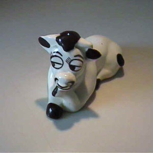 May include: A ceramic cow figurine in a reclining pose. The cow is primarily white with black spots and accents on the ears, horns, and hooves. It has a whimsical, cartoonish design with closed eyes and a smiling mouth.