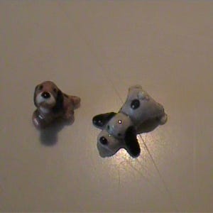 May include: Two small ceramic dog figurines. One figurine is brown, white, and black, depicted in a sitting position. The second figurine is white with black spots and black ears, shown in a lying down position with its head turned toward the side.