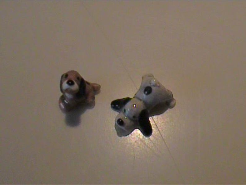 May include: Two small ceramic dog figurines. One figurine is brown, white, and black, depicted in a sitting position. The second figurine is white with black spots and black ears, shown in a lying down position with its head turned toward the side.