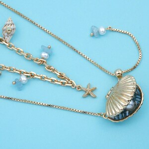May include: Gold-toned necklace with seashell, starfish, and pearl accents. The necklace features a delicate chain design with a seashell pendant and dangling charms. The background is a solid light blue.