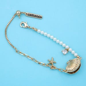 May include: Gold-toned bracelet with a seashell, starfish, and pearl accents. The bracelet features a delicate chain and a clasp closure. The brand name "SUMIYAKI" is displayed on a small tag. The bracelet is set against a light blue background.