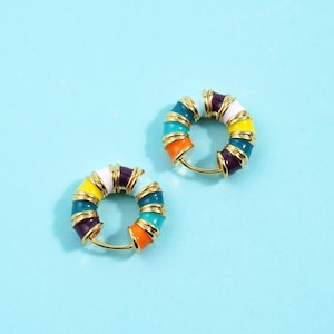 May include: Two colorful hoop earrings. Each earring features a gold-tone metal hoop with a series of vibrant, enameled segments in shades of orange, teal, yellow, white, and purple. The earrings are set against a light blue background.
