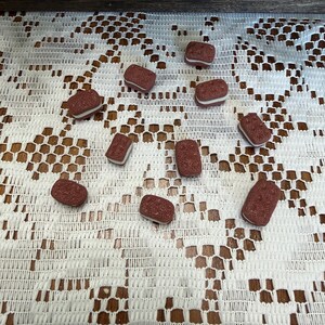 May include: A collection of small, rectangular, reddish-brown objects with a white layer in the middle, scattered on a white lace tablecloth. The objects appear to be a type of candy or treat, possibly resembling a cookie or a similar confection.