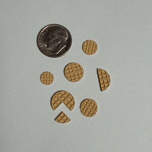May include: A dime next to several miniature, tan-colored waffles of varying sizes and shapes. Some waffles are whole, while others are cut into sections. The waffles have a square grid pattern, and are arranged on a light gray surface.