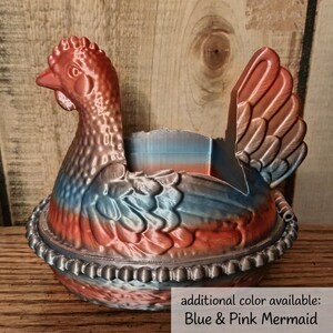 Op de afbeelding: Een decoratieve kippenvormige container in tinten rood, blauw en bruin. De container heeft een gedetailleerd ontwerp, inclusief een kam, vleugels en een kralenrand. De tekst onderaan luidt "additional color available: Blue & Pink Mermaid."