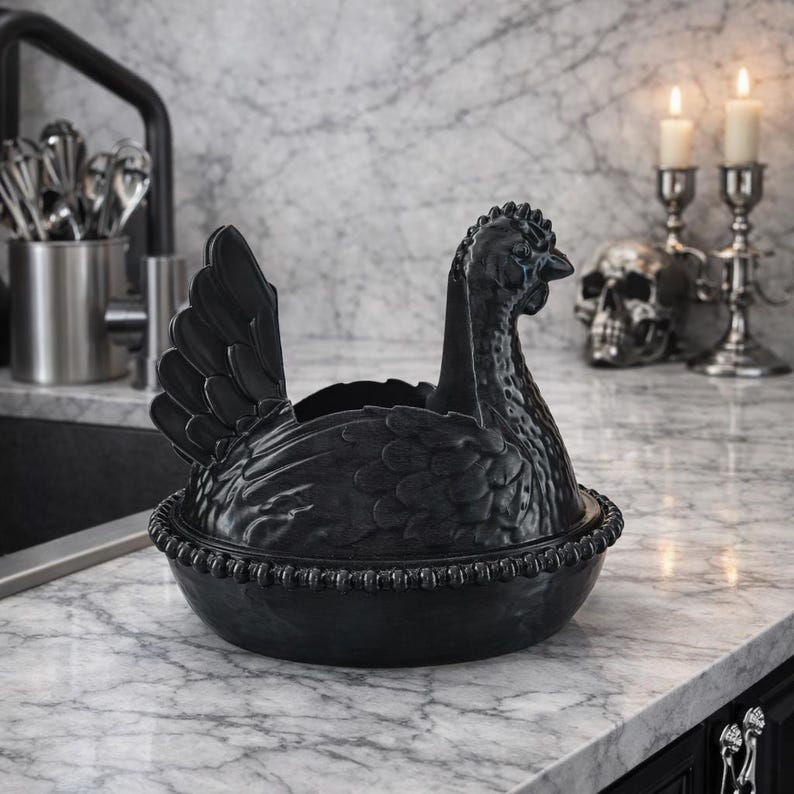 Op de afbeelding: Een zwarte, kippenvormige keramische serveerschaal met een gedetailleerd ontwerp. De schaal heeft een deksel dat het lichaam van de kip vormt en een basis. De kip staat op een marmeren aanrecht. Het is een decoratief keukenartikel.