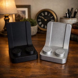 May include: Two LEGO-themed phone stands, one black and one silver, sit on a wooden surface. Each stand has two circular LEGO studs and the LEGO logo. The stands have a curved back and a flat base.