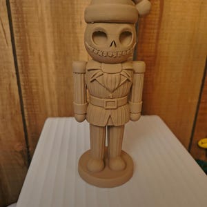 May include: A monochromatic tan nutcracker figurine featuring a skeletal face with hollow eyes and a stitched mouth. The figure wears a Santa hat, a collared jacket with a belt, and stands on a circular base. The entire piece has a matte finish.
