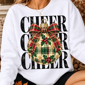 May include: A white long-sleeve sweatshirt featuring the word CHEER repeated three times in a distressed black font. Centered over the text is a large Christmas ornament graphic with a red and green plaid pattern, holly leaves, red berries, and a bow.