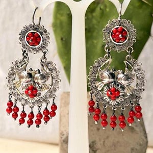May include: A pair of silver dangle earrings featuring a circular filigree frame with two central birds and a cluster of small red beads. The earrings are accented with additional red bead dangles hanging from the bottom of each circular frame.