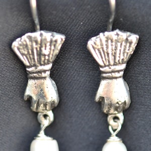 Puede incluir: Un par de pendientes plateados. Cada pendiente presenta un dije en forma de mano que sostiene una perla blanca. Los pendientes tienen un cierre de gancho. El fondo es gris oscuro.