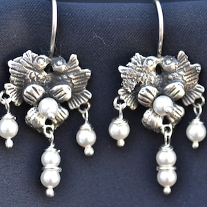 Puede incluir: Un par de pendientes plateados con diseño floral. Cada pendiente presenta un grupo de hojas y flores, con tres perlas falsas colgantes. Los pendientes tienen cierre de gancho y están sobre un fondo azul oscuro.