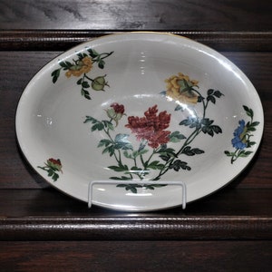 Può includere: Una ciotola ovale bianca con un motivo floreale con fiori rossi, gialli e blu. La ciotola ha un bordo dorato ed è decorata con foglie verdi.