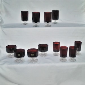 Op de afbeelding: Een verzameling robijnrood glaswerk, waaronder wijnglazen en kleine dessertschalen. De glazen hebben een donkerrode tint en staan op heldere, afgeronde voetjes. De opstelling suggereert een set voor het serveren van drankjes of desserts.