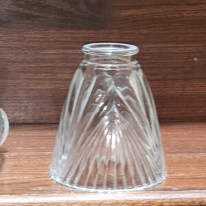 May include: A clear glass light shade featuring a conical shape with vertical ribbed detailing. The shade has a smooth, circular opening at the top and a wider, scalloped base, showcasing a transparent design that allows light to pass through.