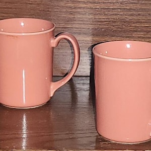 Puede incluir: Cuatro tazas de cerámica rosa a juego con asas curvas. Las tazas están espaciadas uniformemente y se asientan sobre una superficie oscura y reflectante contra un fondo de madera. Las tazas tienen un diseño simple y clásico.