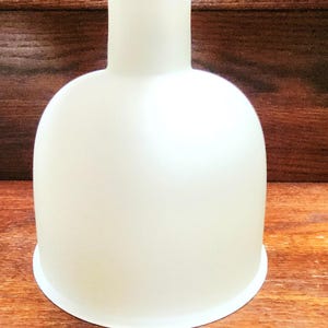 May include: A frosted glass lampshade with a rounded body and a cylindrical neck. The lampshade is opaque and sits on a white base. A ruler is placed inside the neck of the lampshade, showing the number 8.