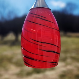 Op de afbeelding: Een rode glazen hanglamp met zwart wervelontwerp. De lamp heeft een zilverkleurige metalen bovenkant en een druppelvorm. De achtergrond is wazig en toont een natuurlijke buitenomgeving.
