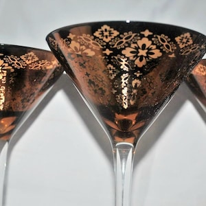 May include: Three vintage martini glasses with a black and gold floral design. The glasses have a classic triangular bowl shape and a long, clear stem. The gold pattern is intricate and covers the rim and bowl.
