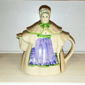 May include: A ceramic teapot shaped like a person wearing a bonnet with purple flowers, a cream-colored cape with green trim, and a purple apron. The figure has a painted face, a handle on the right side, and a spout on the left side of the body.