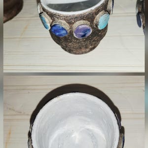 May include: A small, handcrafted ceramic pot with a brown exterior and a white interior. The pot is adorned with decorative, colorful, circular accents in shades of blue and purple. The pot is likely used for holding small items.