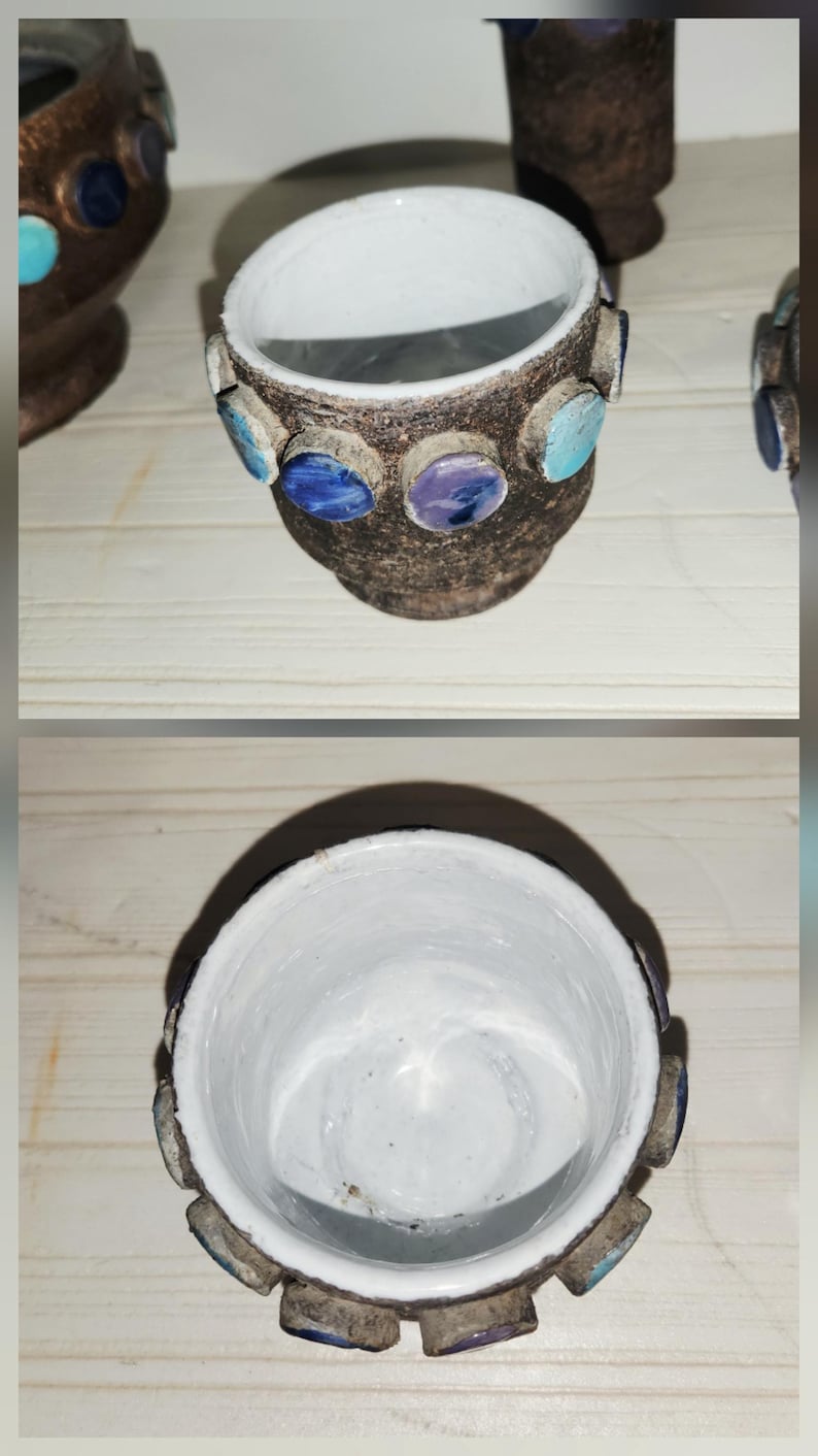 May include: A small, handcrafted ceramic pot with a brown exterior and a white interior. The pot is adorned with decorative, colorful, circular accents in shades of blue and purple. The pot is likely used for holding small items.