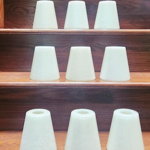 Op de afbeelding: Negen crèmekleurige, kegelvormige lampenkappen zijn gerangschikt op houten planken. De kappen hebben een matte uitstraling en een licht gestructureerd oppervlak. De kappen zijn gelijkmatig verdeeld en lijken van glas of een vergelijkbaar materiaal te zijn. De achtergrond is een warm, middentoon hout.