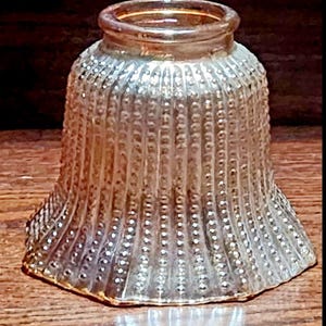 May include: A clear, bell-shaped glass lampshade with a textured, dotted pattern. The shade has a flared base and a slightly rounded top, reflecting light. The glass has a vintage, antique appearance.
