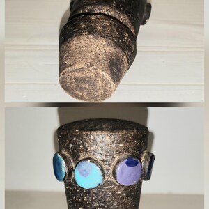 May include: A handcrafted, dark brown ceramic vase with a textured surface. The vase is adorned with three circular, glazed accents in blue and purple. The vase has a cylindrical shape with a slightly flared base.