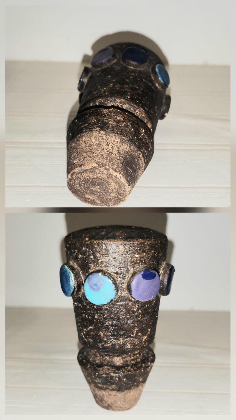 May include: A handcrafted, dark brown ceramic vase with a textured surface. The vase is adorned with three circular, glazed accents in blue and purple. The vase has a cylindrical shape with a slightly flared base.