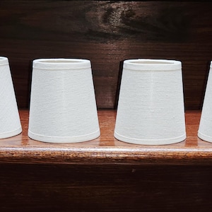 May include: A set of four small, white, tapered lamp shades with a subtle, fine-textured fabric finish. Each shade features a clean, cylindrical shape with a slightly wider base and a narrower top, designed for clip-on or small lamp fixtures.