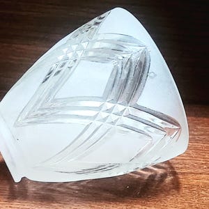Puede incluir: Un objeto decorativo de vidrio esmerilado con un patrón geométrico de diamantes. El objeto tiene forma triangular con base plana y descansa sobre una superficie de madera. El vidrio tiene aspecto esmerilado y el patrón de diamantes está grabado.