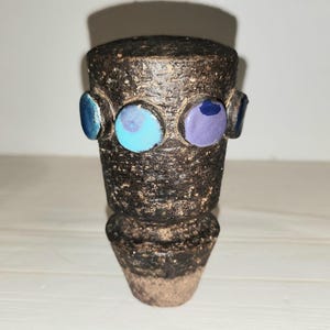 May include: A handcrafted, dark brown ceramic vase with a textured surface. The vase features three colorful, circular accents in shades of blue and purple. The vase has a cylindrical body and a flared base, showcasing a unique, artistic design.