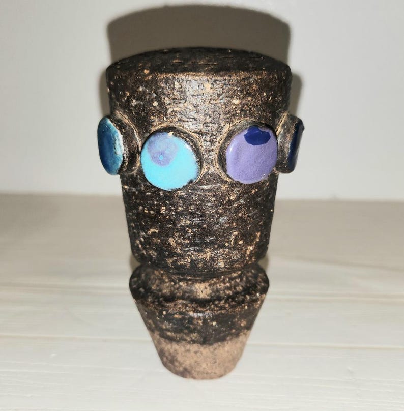 May include: A handcrafted, dark brown ceramic vase with a textured surface. The vase features three colorful, circular accents in shades of blue and purple. The vase has a cylindrical body and a flared base, showcasing a unique, artistic design.