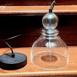 May include: A clear glass pendant light fixture with a bell-shaped shade and a round top. The fixture has a black wire and a black circular base. The light is designed to be suspended from the ceiling.