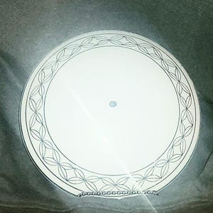 May include: A white, round decorative plate with a detailed, geometric border design in gray. The plate has a small, light blue dot in the center. The plate is resting on a gray surface.