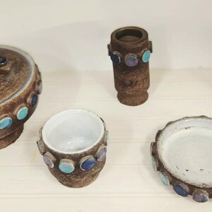 May include: A collection of brown ceramic pottery pieces, including a lidded jar, a small vase, a cup, a dish, and a cylindrical container. Each piece is adorned with turquoise and purple circular accents.