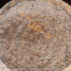 May include: Close-up of the bottom of a weathered, tan-colored ceramic object. The surface is textured with a circular design and what appears to be a partial inscription in a lighter shade of brown. The object shows signs of age and wear.