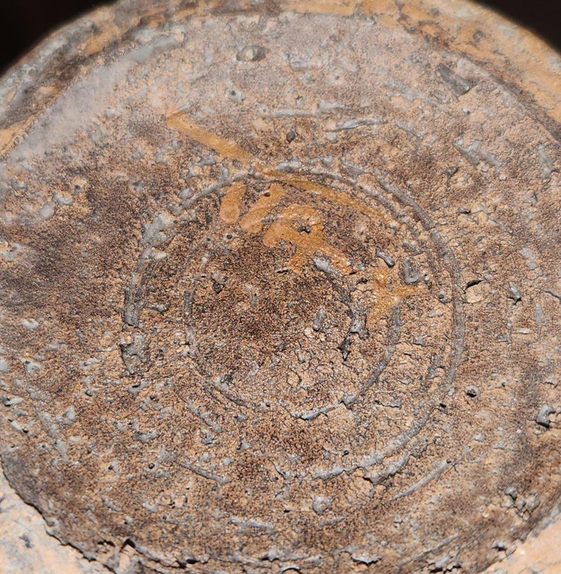 May include: Close-up of the bottom of a weathered, tan-colored ceramic object. The surface is textured with a circular design and what appears to be a partial inscription in a lighter shade of brown. The object shows signs of age and wear.
