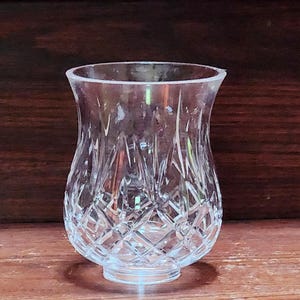 Puede incluir: Un portavelas votivo de cristal tallado transparente. El cristal tiene un patrón de diamantes en la parte inferior y un borde liso y acampanado. El portavelas se asienta sobre una pequeña base circular. El fondo es una superficie de madera marrón oscuro.