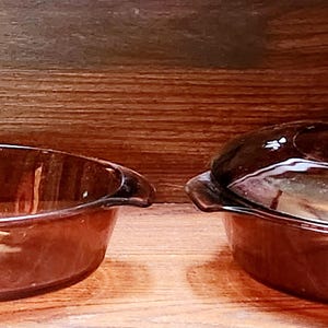 Puede incluir: Dos fuentes para hornear de vidrio marrón con tapas. La fuente de la izquierda es redonda y la fuente de la derecha es ovalada.