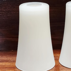 Op de afbeelding: Twee crèmekleurige, kegelvormige lampenkappen. De kappen hebben een gladde, matte afwerking en een licht uitlopend ontwerp. Ze staan tegen een donkere houten achtergrond.