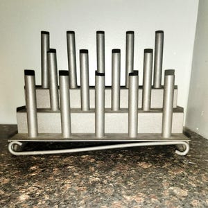 Pode incluir: Um suporte de utensílios de metal prateado com vários postes verticais, cada um encimado por uma tampa preta. O suporte tem uma base retangular e uma alça de arame curvada. O item foi projetado para guardar utensílios de cozinha.