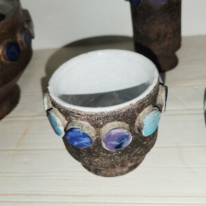 May include: A small, handcrafted ceramic pot with a dark brown exterior and a white interior. The pot is adorned with colorful, circular accents in shades of blue, turquoise, and purple. The pot is likely used for decorative purposes.